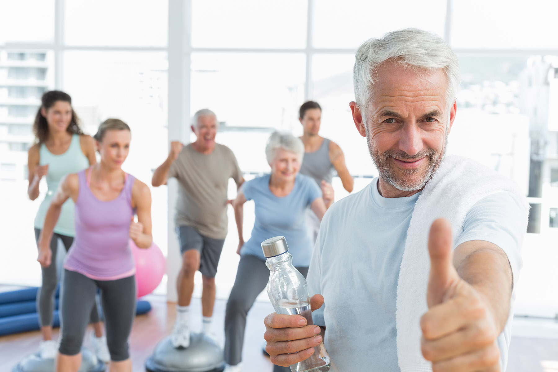 Happy senior man gesturing thumbs up with people exercising in the background at fitness studio